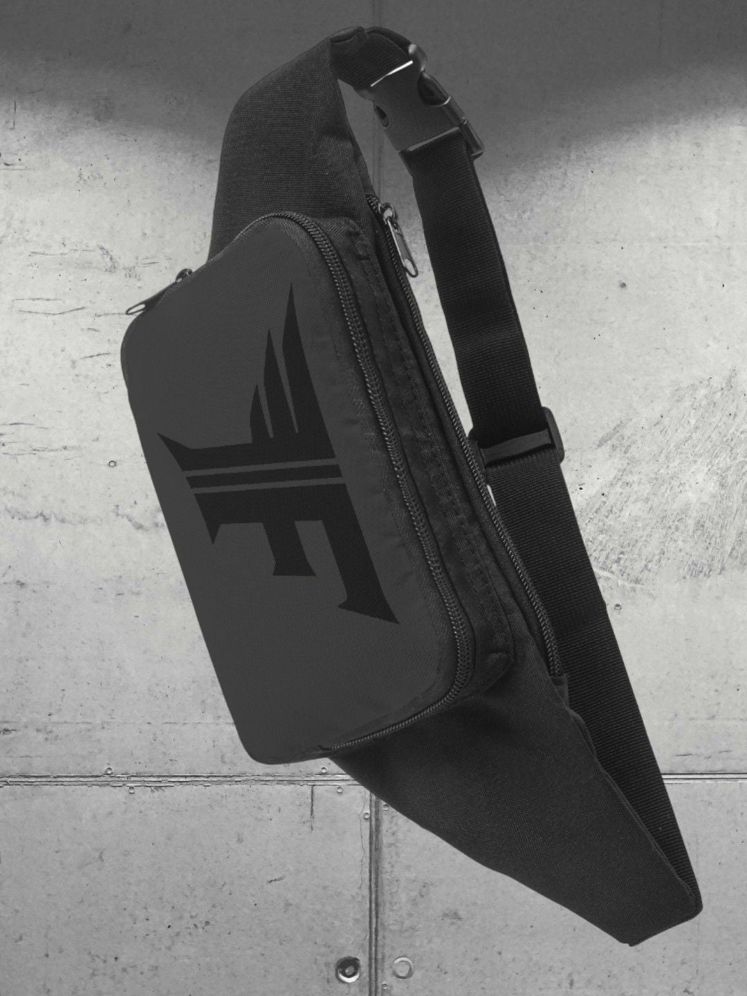 A black fanny pack/sling bag with a dark, prominent stylized 'F' logo hanging from its strap against a background of light gray concrete or metal panels.