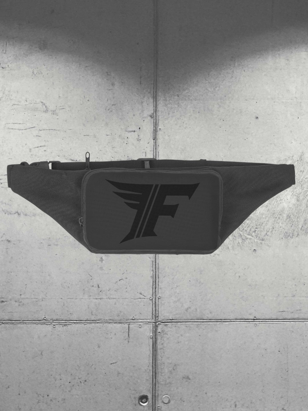A black fanny pack is centered against a gray background of metal panels. The pack features a prominent, dark, stylized 'F' logo on the front pouch.